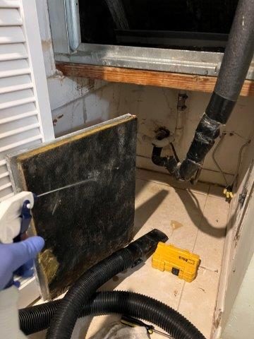 A gloved hand sprays a dark-stained filter panel next to black drainage hoses, a taped pipe, and a yellow toolbox under a window.