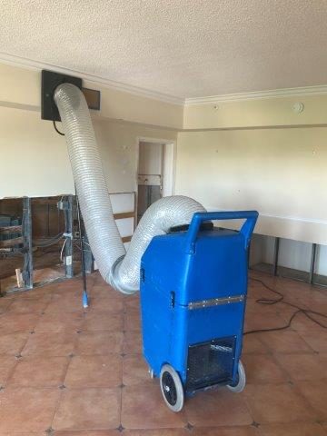 A blue commercial drying unit with a flexible duct is connected through a wall vent in a tiled interior room.